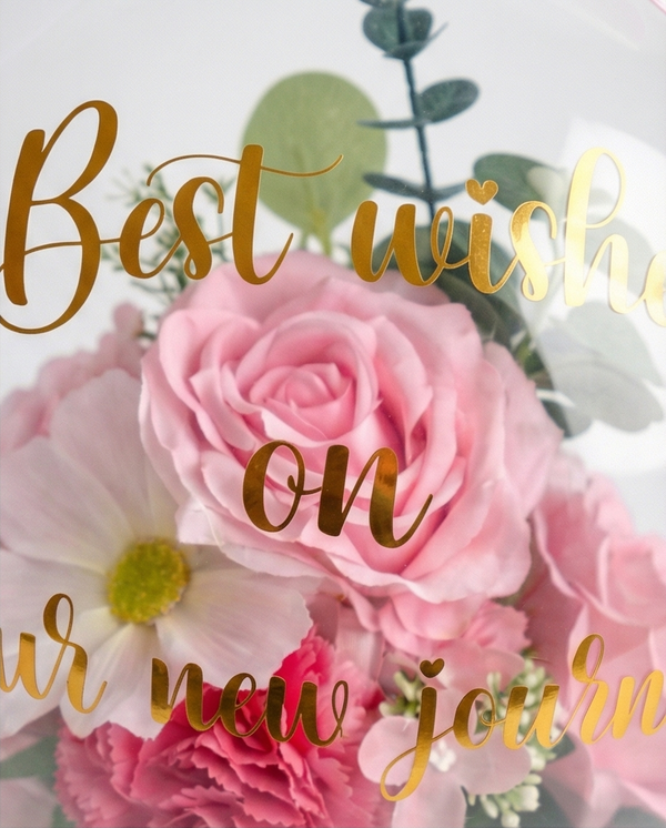 Pink floral arrangement in a transparent dome with 'Best wishes on your new journey' text, text close up