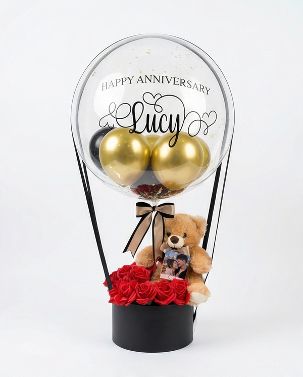 Decorative balloon with gold balloons, teddy bear, and red roses inside a clear dome, full view